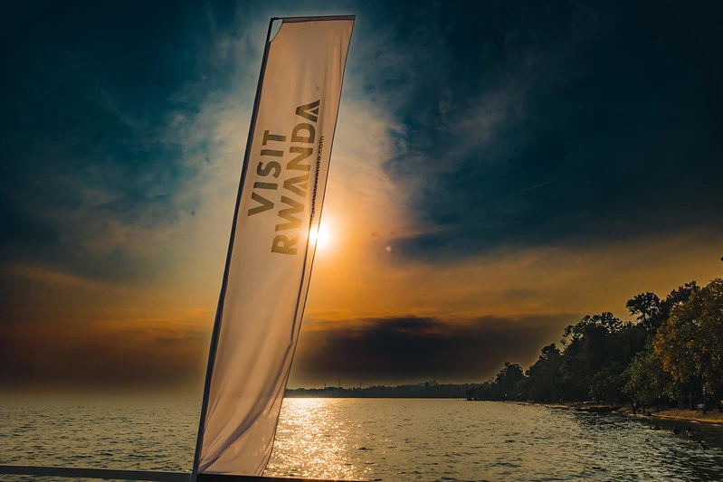 Visit Rwanda flag displayed by the shores of Lake Kivu, with calm waters and scenic hills in the background.